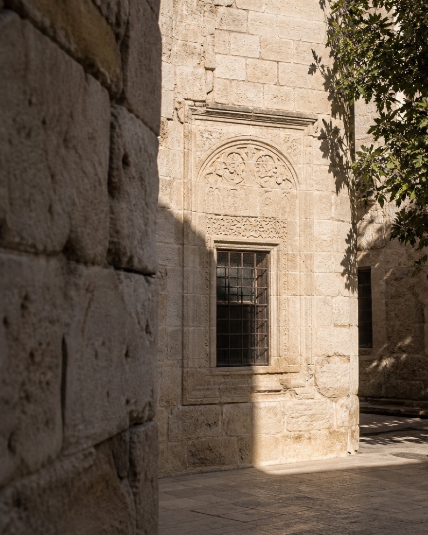 Stone courtyard wall with a framed grated window and carved arch in sharp light and shadow