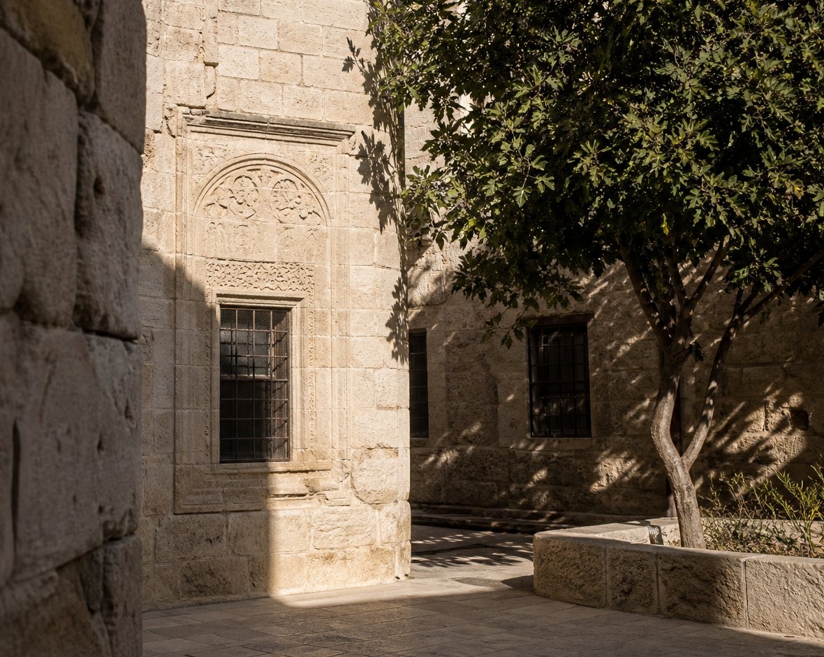 Sunlit stone courtyard, a grated window and a tree against a quiet facade